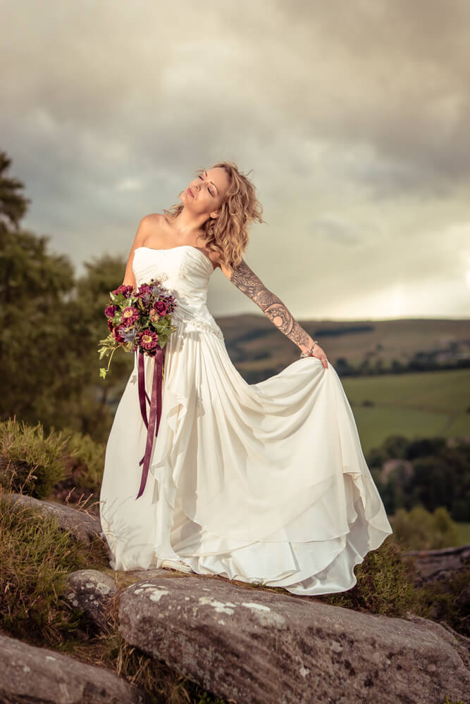 A bride stands on a rock during a Peak District Post-Wedding Shoot holding her skirts out to the wind as the Derbyshire landscape is pictured in the background
