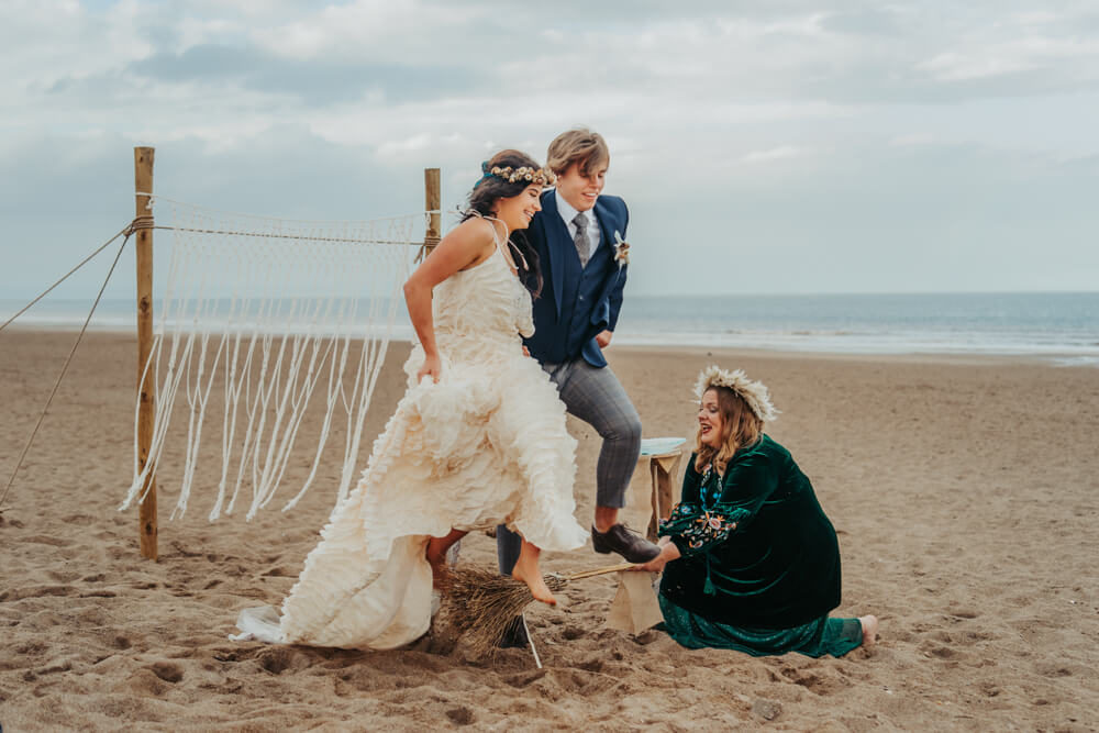 A bride and groom jump over a broom held by a celebrant on their eco-friendly beach elopement