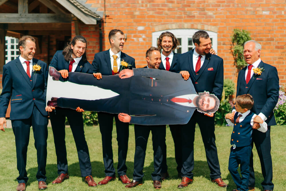 Groomsmen laugh as they hold up a life-size cardboard cutout of the groom's brother who couldn't attend the wedding at Donington Park Farmhouse