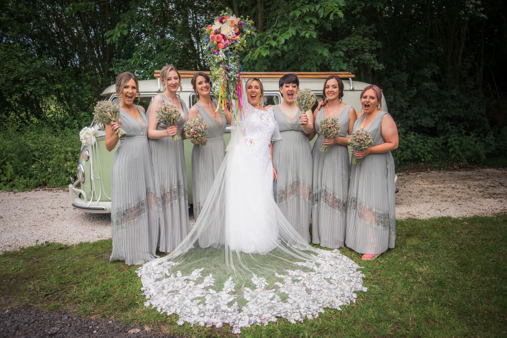 A bride thrusts her colourful bouquet in the air as she stands with her veil spread in front of her, accompanied by her six bridesmaids in olive green in front of a green VW campervan at Hidden Hive tipi wedding venue