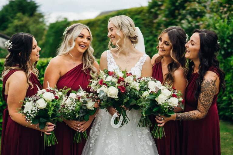 Bride laughs with her four bridesmaids who are dressed in red as they hold their bouquets together and pose for a picture in the gardens at Melbourne View Hotel