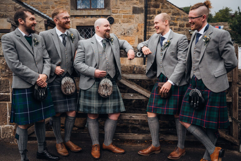 Groom and his four groomsmen are wearing kilts and leaning on a gate before their home garden ceremony in Derbyshire