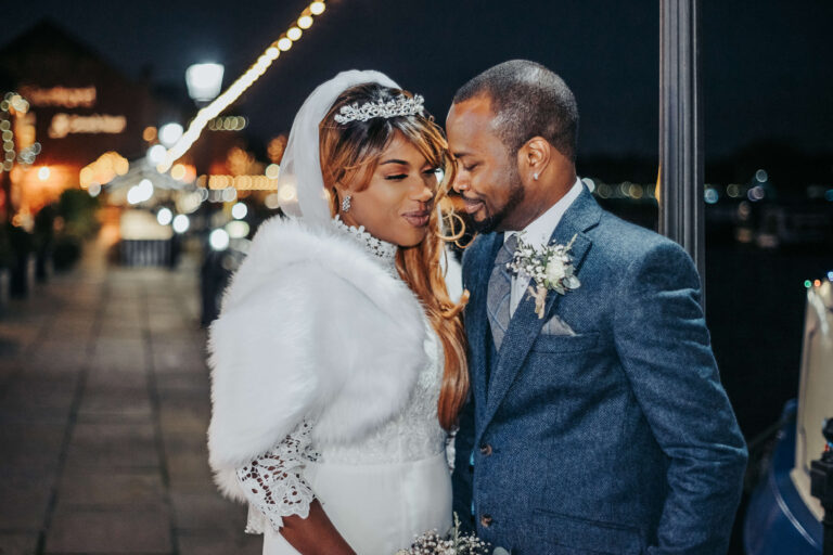 A newlywed couple stand close together in the winter night with the bokeh lights of Mercia Marina behind them