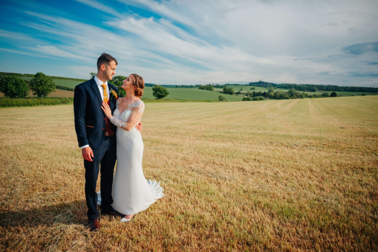 A young couple pose in a golden field near Donington Park Farmhouse with the Derbyshire landscape and blue skies behind them.