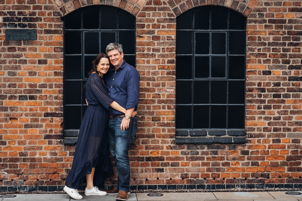 An engaged couple, both in blue, cuddle next to a pair of arched windows and an industrial style brick wall at The Silk Mill in Derby for their engagement shoot