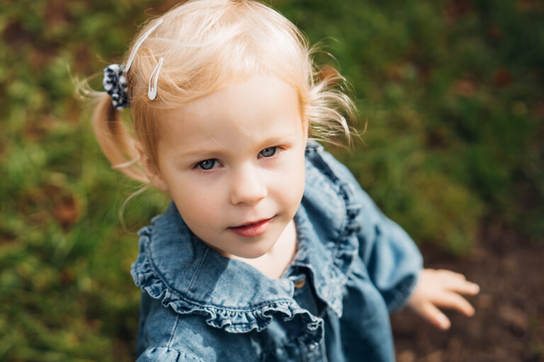 A pretty blonde two-year-old girl looks up at the camera wearing a denim dress with grass in the background