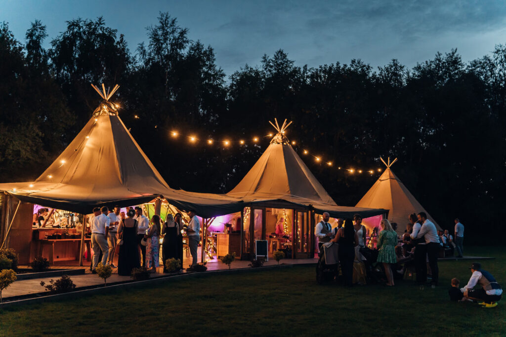 The three tipis of The Hidden Hive in Derbyshire shown lit up at night with festoon lighting and guests partying