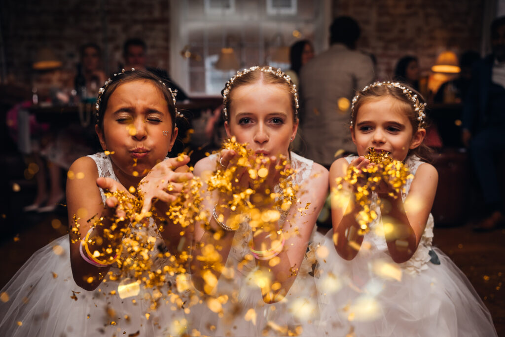 Three flowergirls blow gold confetti at the camera at the West Mill Derby
