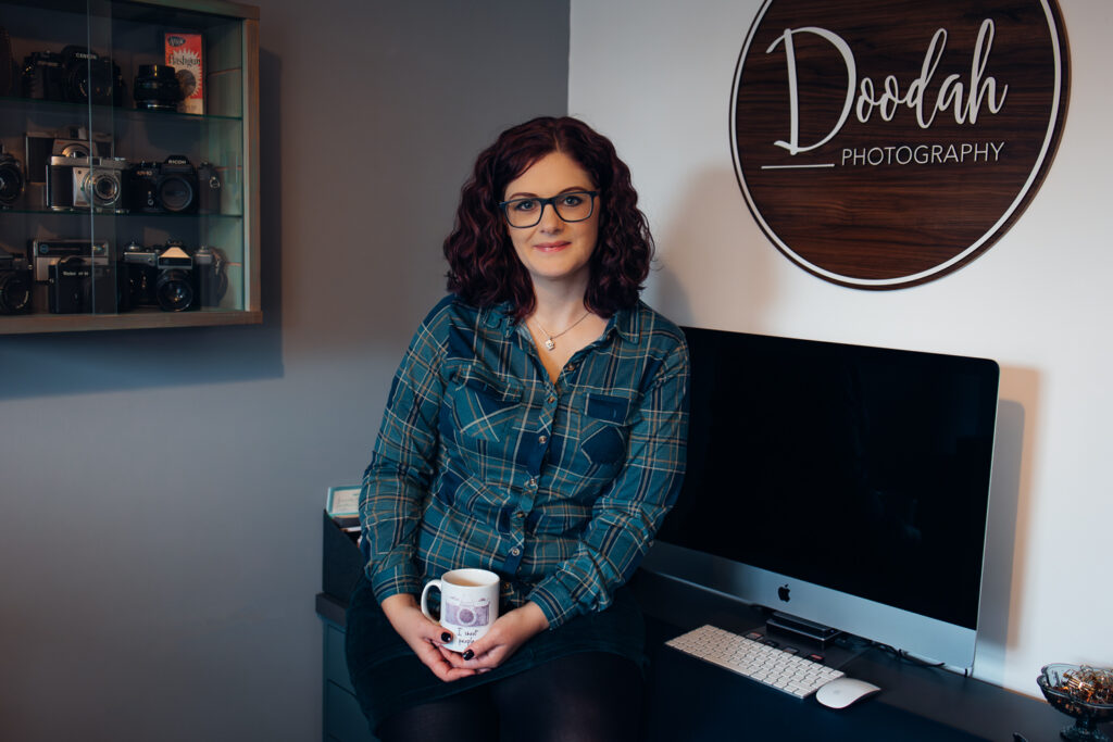 Wedding photographer Emma Duder sitting on the desk in her home office with a mug of tea in front of a Doodah Photography sign