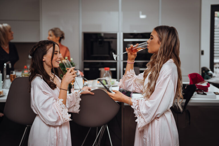 Two teenage bridesmaids face each other and drink champagne whilst getting ready photos are taken